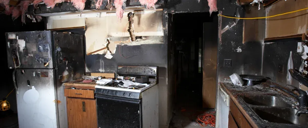 A severely fire-damaged kitchen showing charred walls, appliances, a stovetop, and sink, with insulation hanging from the ceiling.