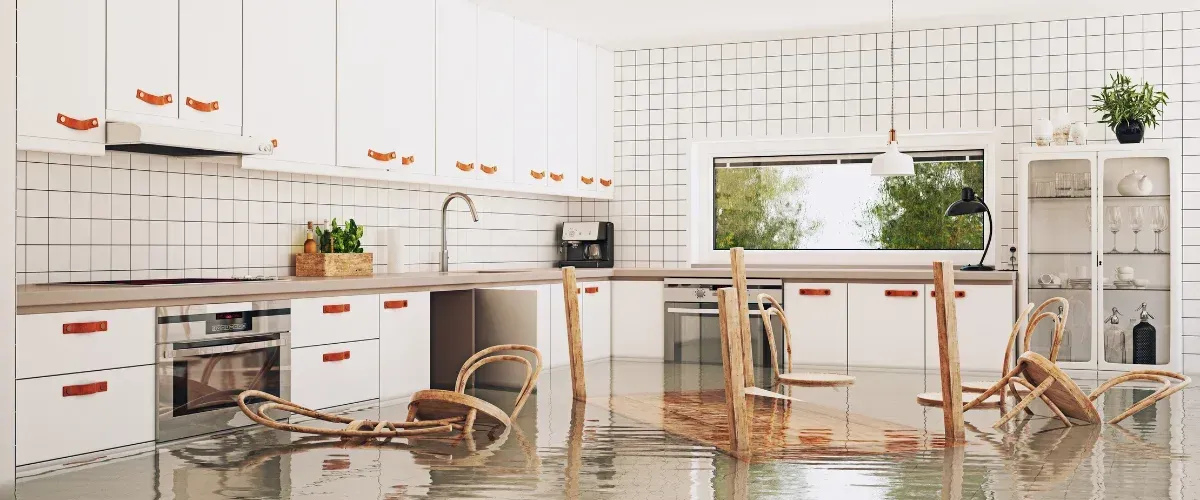 A modern white kitchen is shown submerged in floodwater with wooden chairs floating and toppled over.