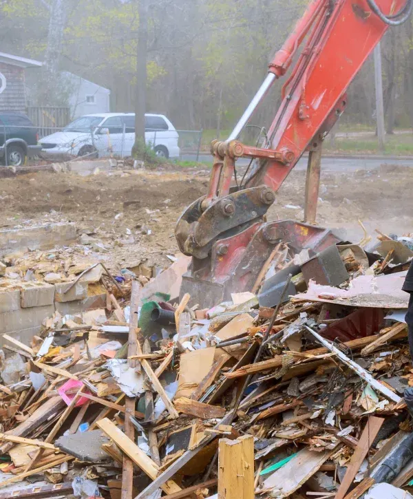 A red excavator's arm and bucket reaching into a large pile of wooden and concrete demolition debris.
