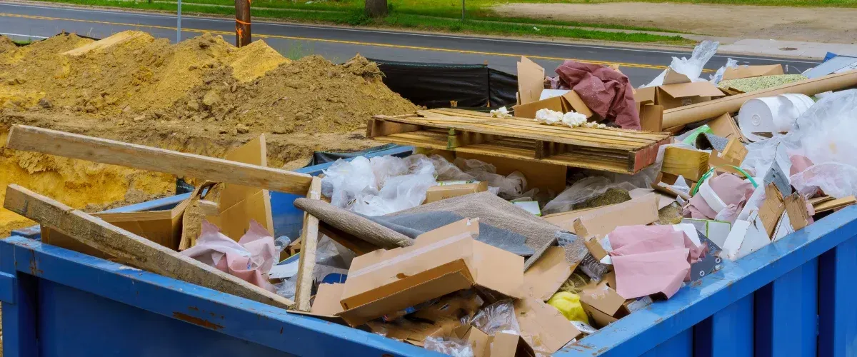 An overflowing blue construction dumpster filled with cardboard, wood, plastic, and debris next to a pile of dirt.
