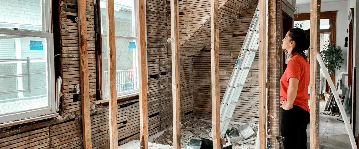 A woman in a red shirt inspects a home renovation project with exposed studs and lath walls.