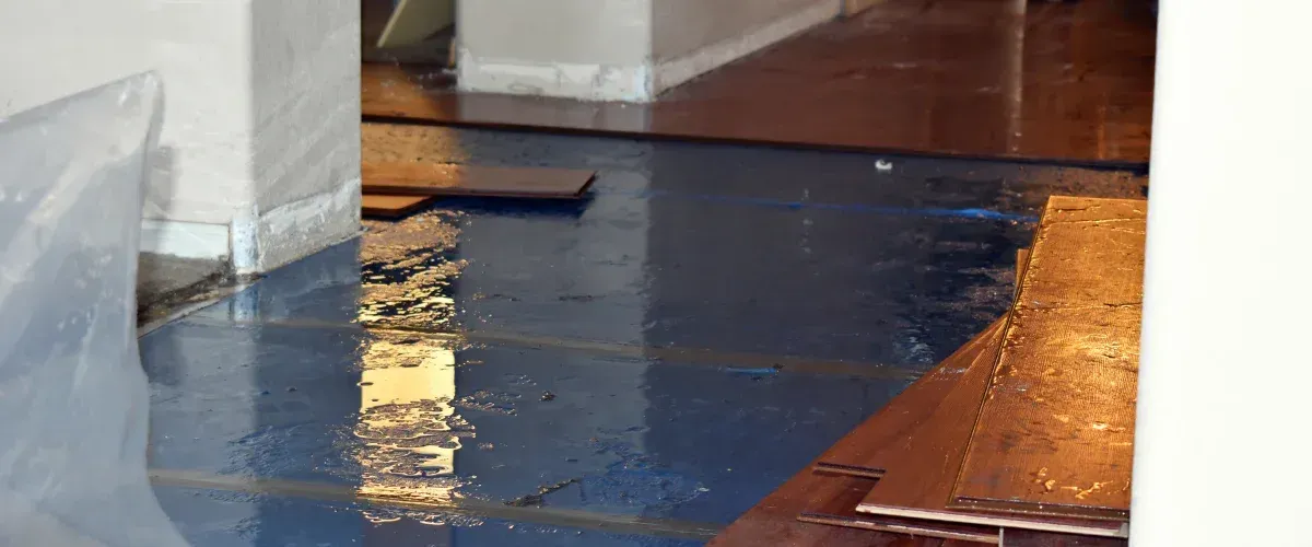 A close-up view of a wet floor with dark blue underlayment and several stacked wooden planks that appear water-damaged.