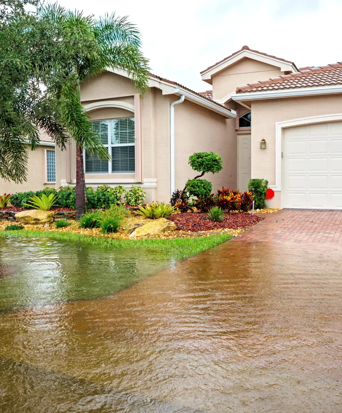 A beige stucco house with a terracotta roof and tropical landscaping, with its brick driveway and lawn partially flooded.