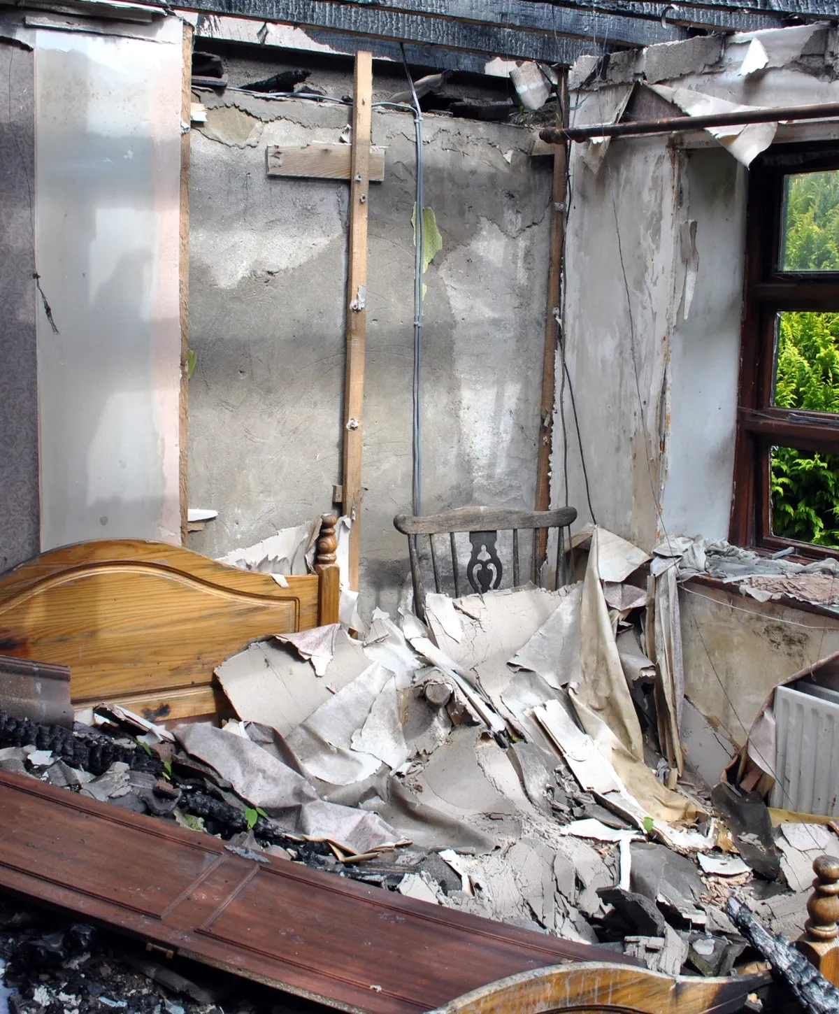 A severely damaged room interior shows a charred ceiling, crumbling walls, and debris-filled floor with parts of a wooden bed and chair.
