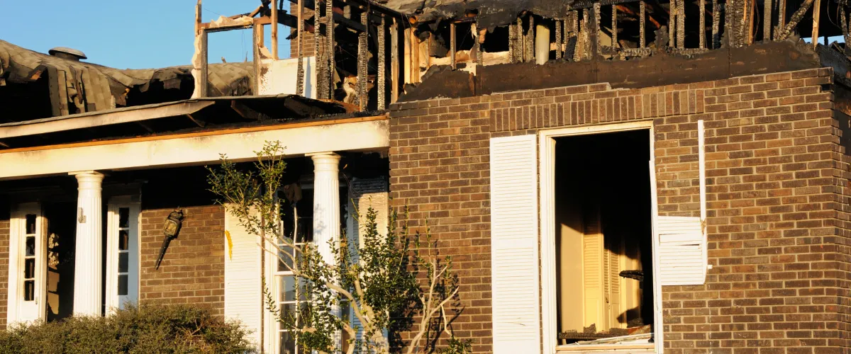 A two-story brick house with a severely burnt and collapsed roof, exposing charred wooden beams against a blue sky.