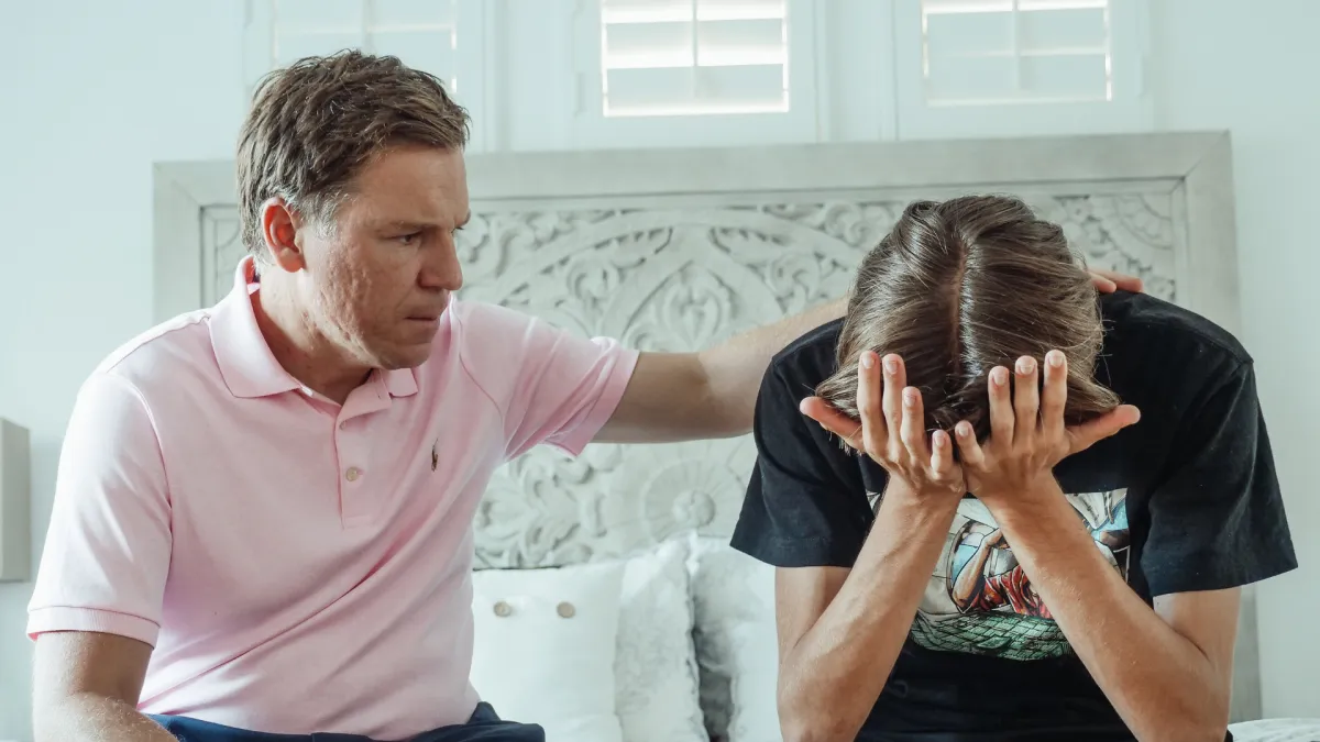 A father comforting his distressed teenage son who is sitting with his head in his hands, illustrating parental support for an anxious child.