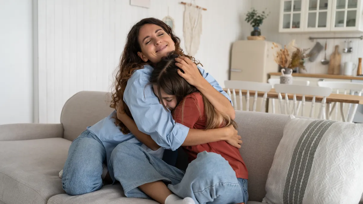 Mother gently hugging her upset daughter on a couch at home, offering comfort, emotional support and a sense of safety during an anxious moment.