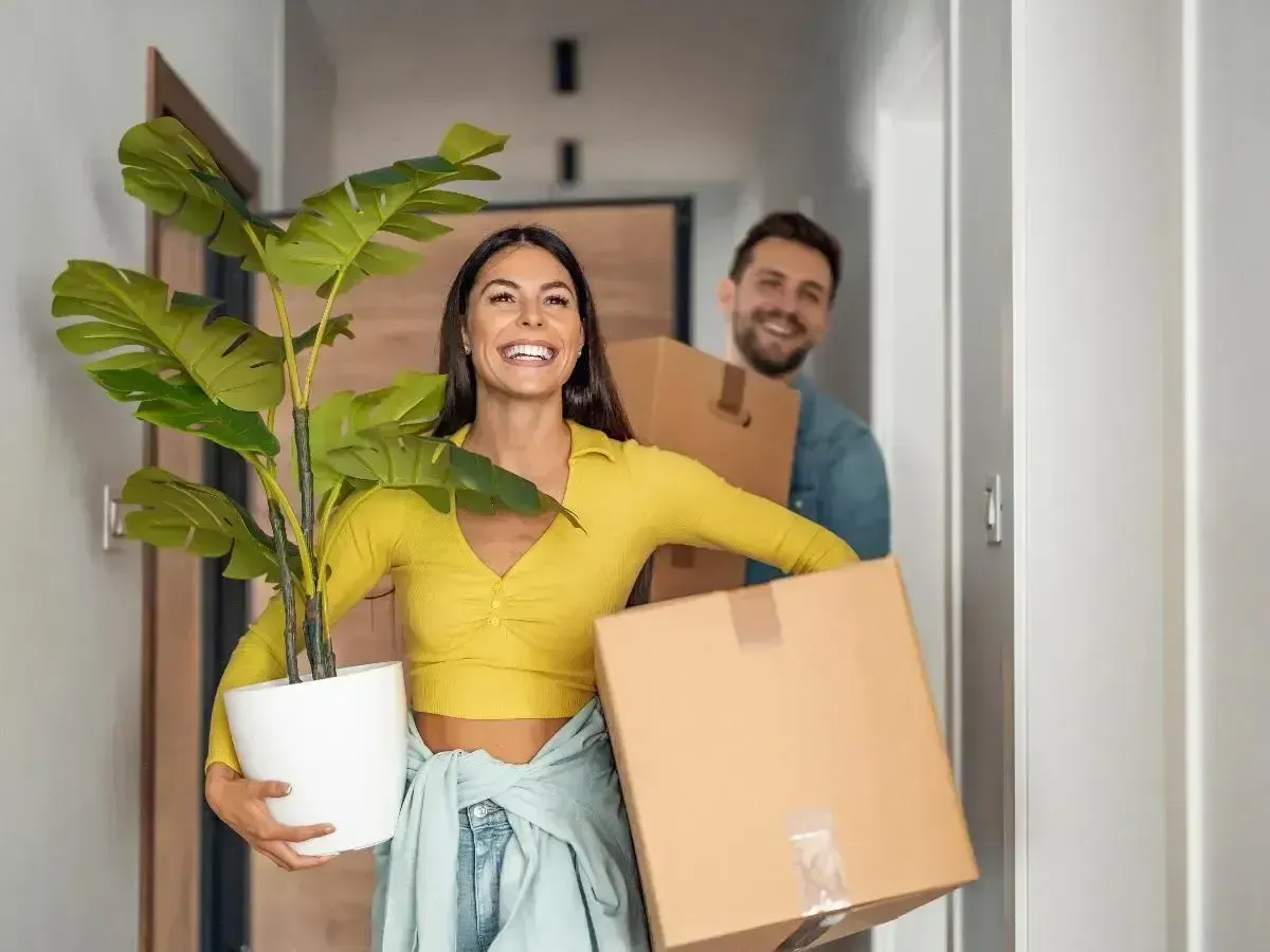 Happy couple moving into their new Galveston apartment, carrying boxes and a plant.