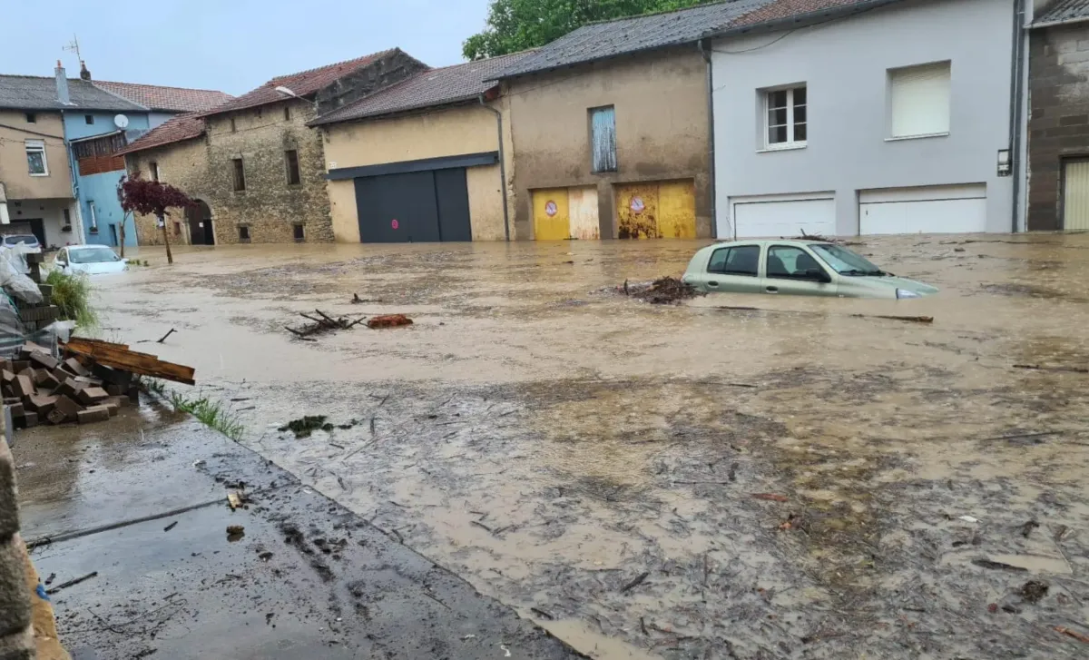 Rue inondée avec des maisons et des garages submergés par l'eau, des débris flottants, et une voiture immergée partiellement dans une zone résidentielle, illustrant les dégâts causés par une inondation.