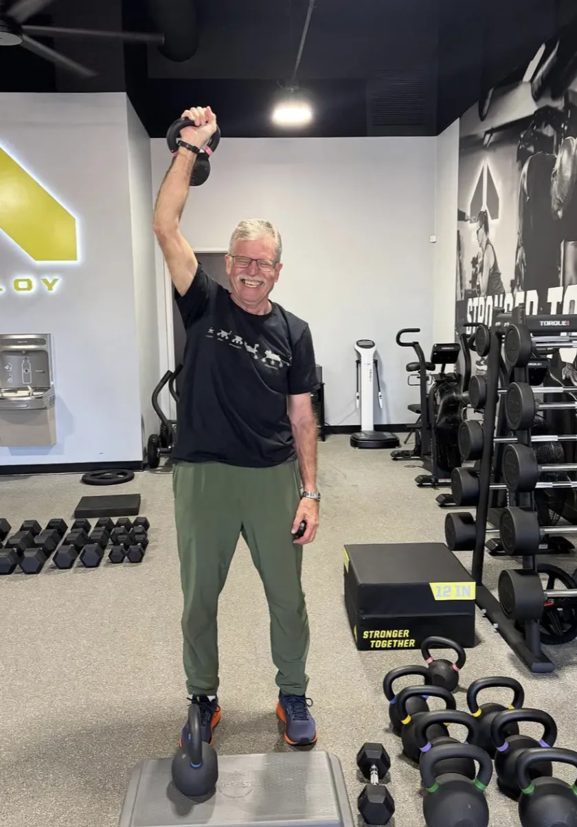 Member smiling and lifting a kettlebell during a strength training session at Alloy Personal Training in Parker, CO. The gym is equipped with kettlebells, dumbbells, and workout equipment designed for full body fitness