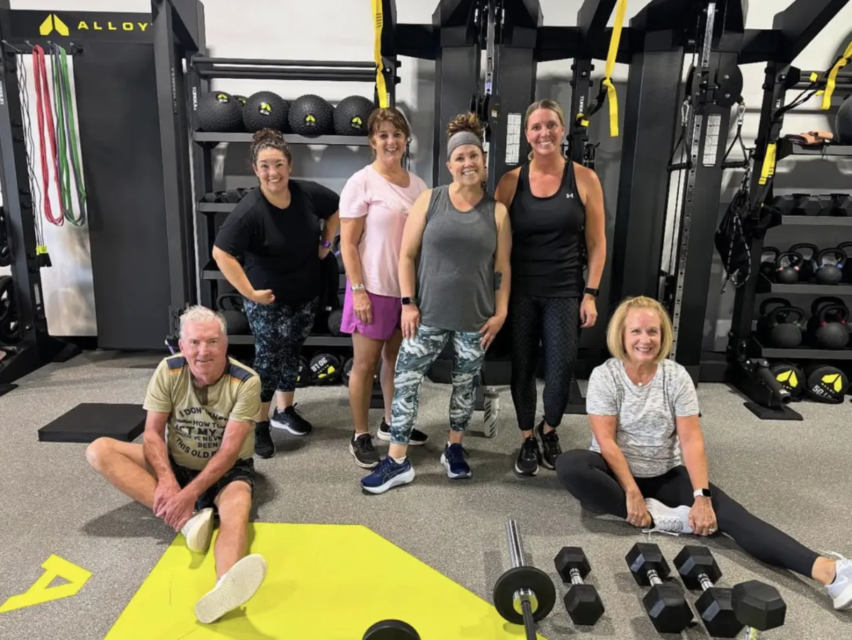 A group of six members in a small-group fitness class at Alloy Personal Training in Parker, CO, after a workout. The diverse group is smiling and celebrating their fitness achievements in a motivating environment
