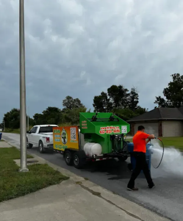 Worker pressure washing trash bin using a professional truck-mounted cleaning system in a residential neighborhood.
