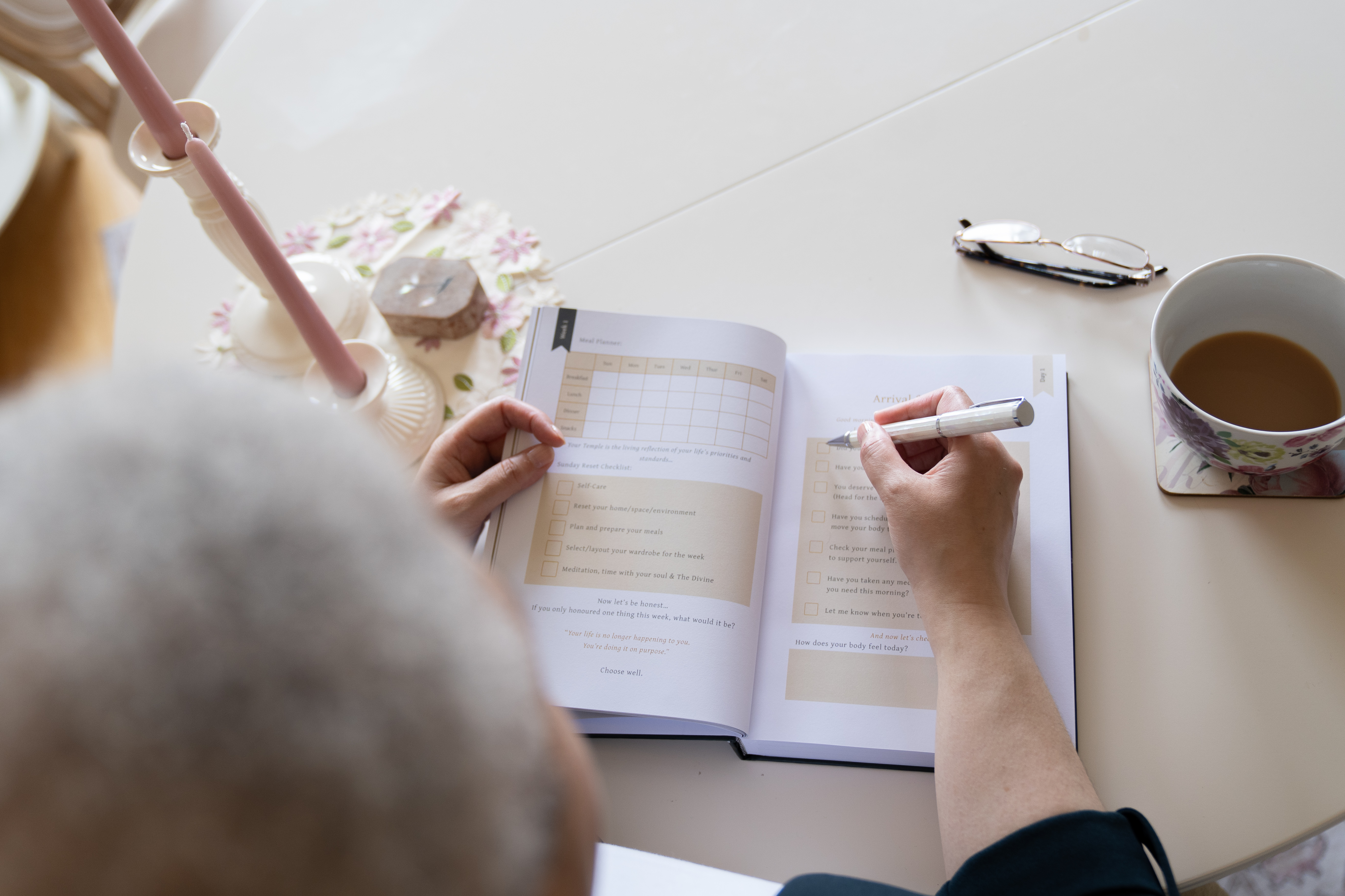 Overhead view of a woman using The Life Companion Journal beside a cup of tea, with the journal's guided pages open.