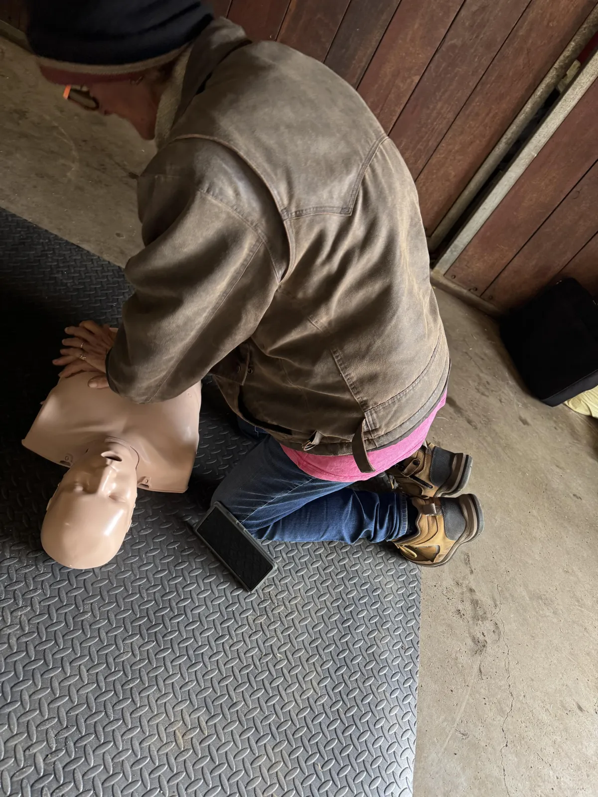 Construction worker in high-vis PPE performing CPR on a training manikin at a worksite, with an open first aid kit nearby