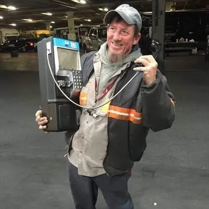 A calm, professional First Aid trainer with a gentle smile, demonstrating the use of a modern first aid kit in a bright, welcoming classroom. The trainer is wearing a branded polo shirt, and diverse adult learners are visible in the background, all engaged and attentive.