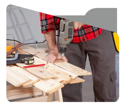 A handyman in a red plaid shirt using a drill to secure wooden planks on a workbench, with tools like a measuring tape and pencil nearby.