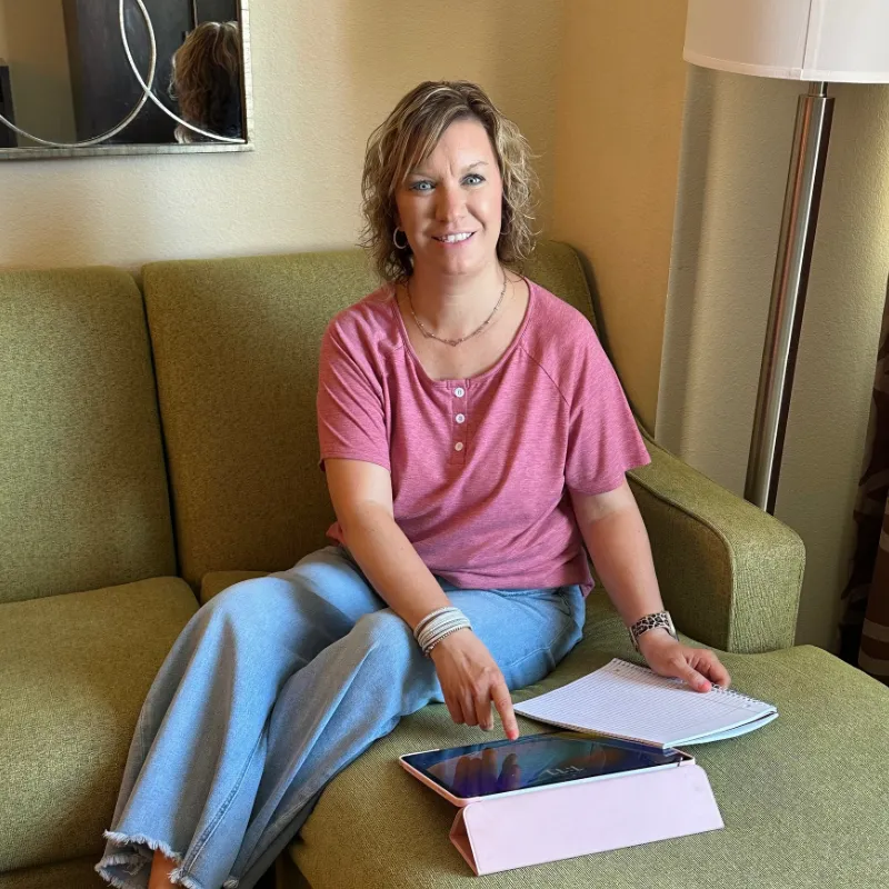 Christian mom sitting on a couch using a tablet and notebook to plan her evening routine for a calm, organized home and business. 
