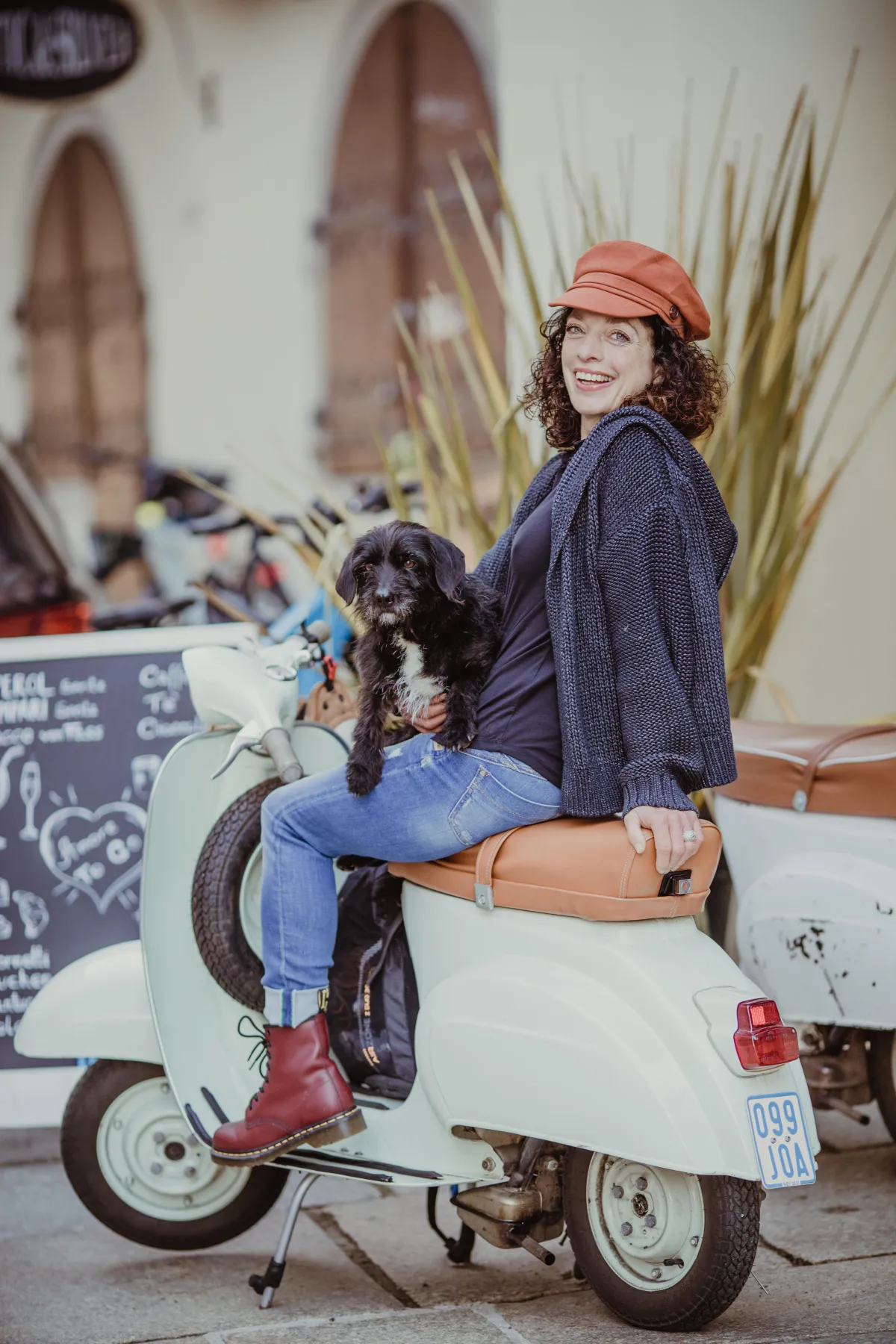 Lifestyle Branding Shooting: Kreative Frau sitzt mit ihrem Hund auf einer weißen Vintage-Vespa vor einem gemütlichen Straßencafé.