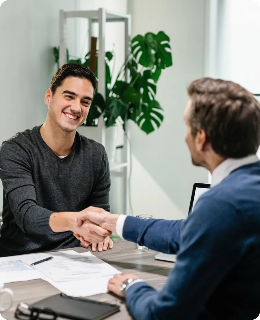 Two men shaking hands over a table in a bright room.