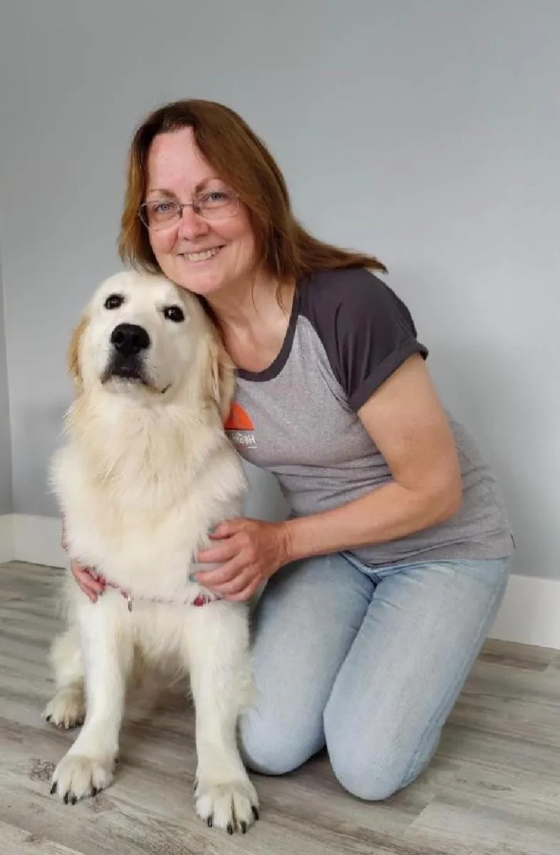 A professional portrait of Alexis Monroe, a woman in her early 40s with curly brown hair and warm smile, wearing a tailored blue blazer and holding a small dog. The background is a softly lit studio with subtle pet motifs, conveying expertise and approachability.