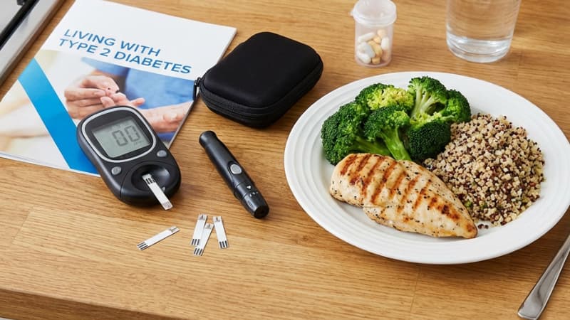 Blood glucose meter with test strips beside a healthy plate of grilled chicken, broccoli, and quinoa with a Type 2 diabetes management guide.