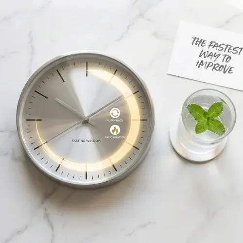 fasting window clock rests on a white marble surface next to a glass of water with mint and a note reading "THE FASTEST WAY TO IMPROVE."