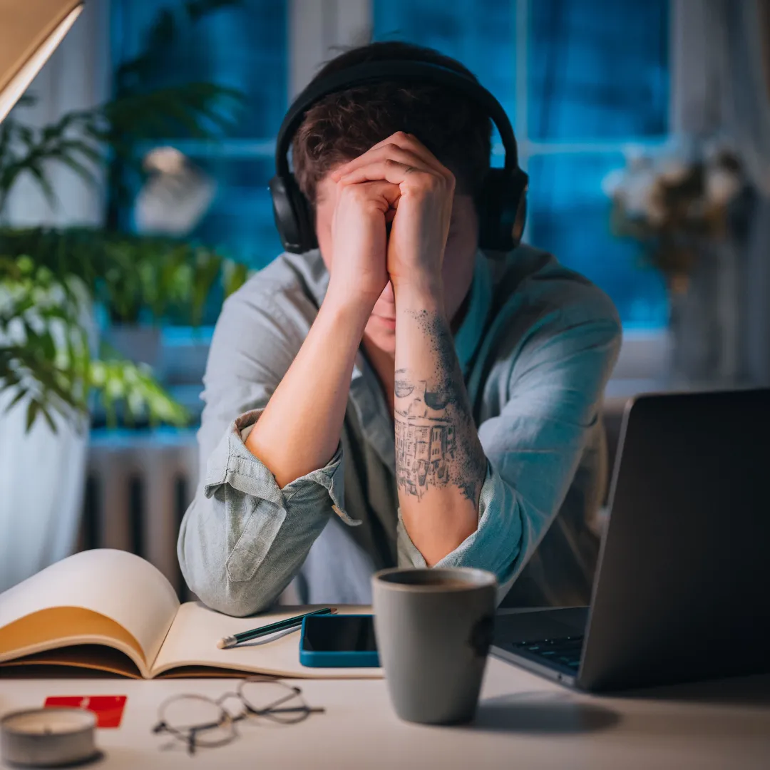 Illustration of a cluttered desk at dusk with crumpled sketches, a dim lamp, and a laptop showing a paused design app; warm muted palette capturing overwhelm and hesitation.