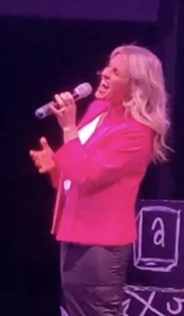 A confident female singer in her twenties, mid-performance, reaching a powerful high note on stage with dramatic lighting and a cheering audience in the background. The scene captures energy, focus, and the thrill of live performance.