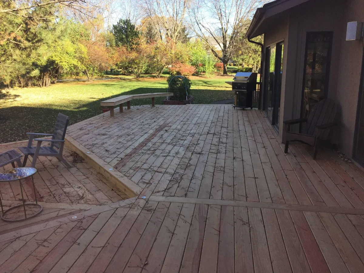 A spacious wooden deck with chairs and a table next to a house, overlooking a grassy backyard with trees. patio extends to a barbecue grill on the right side.