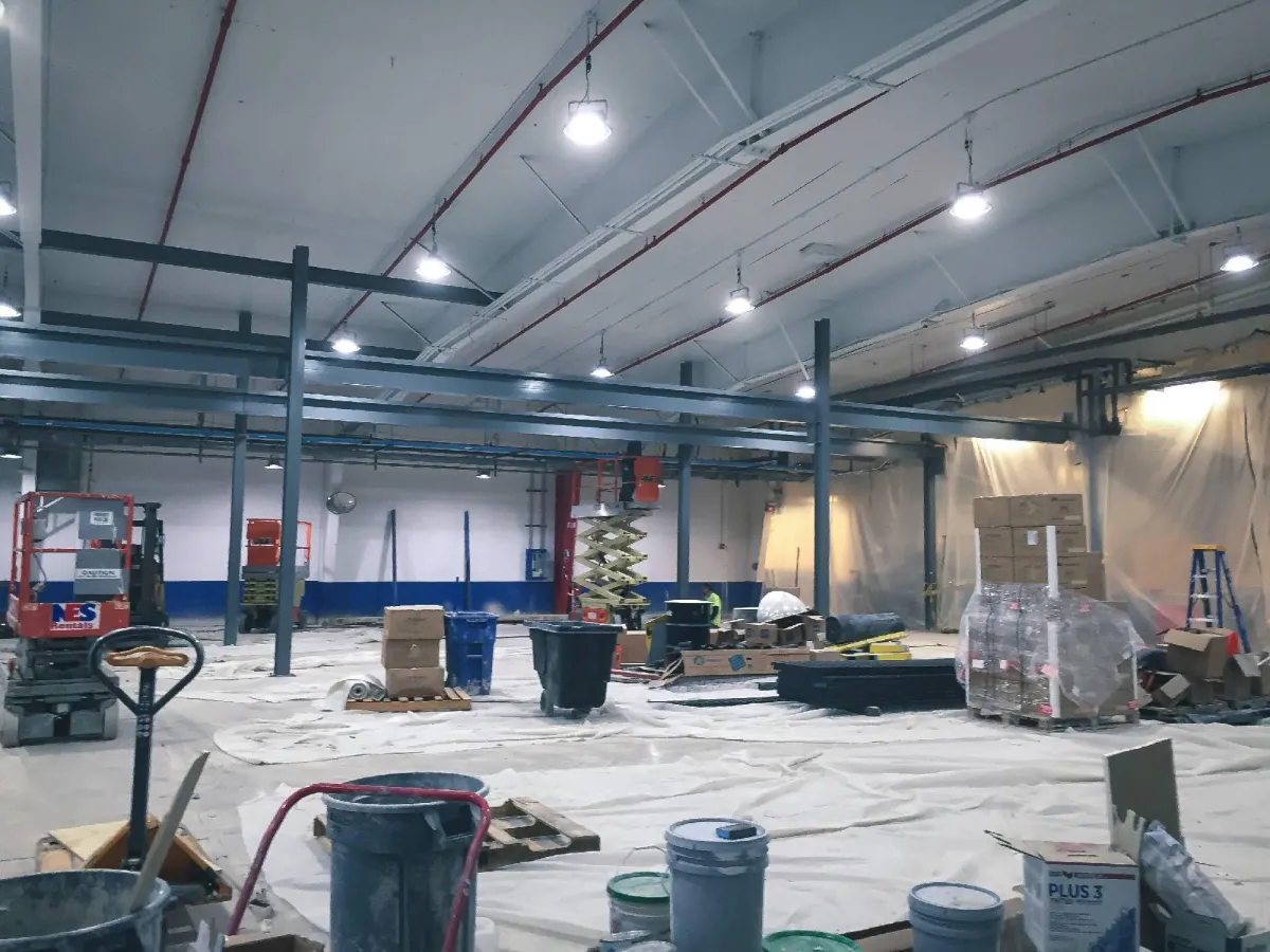 Interior of a spacious industrial construction site showing ongoing work with various construction materials, tools scattered around, and machinery such as a scissor lift. protective sheets hang in the background.