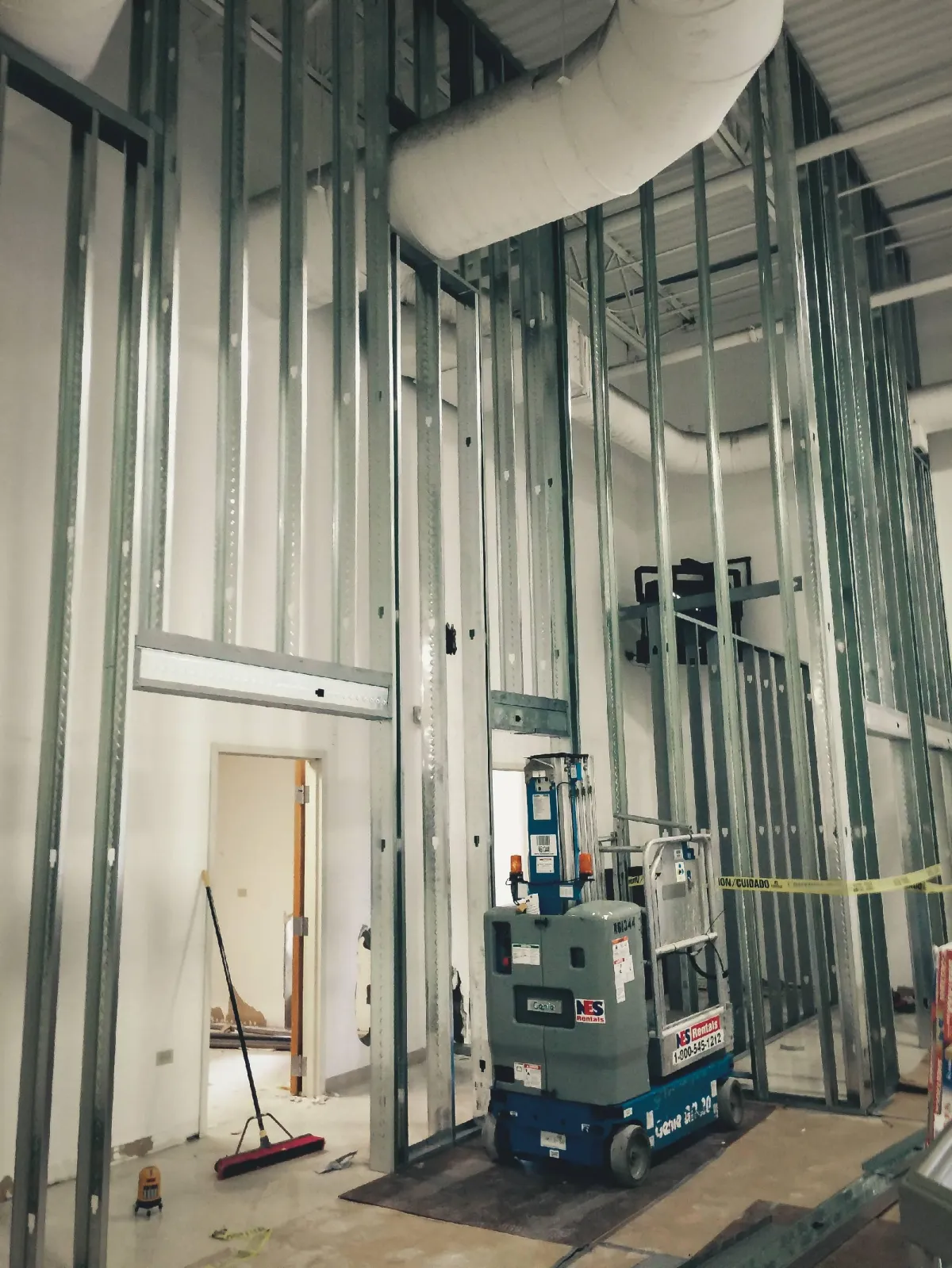 Interior construction site with metal stud framing and a scissor lift, ductwork overhead, and scattered tools in a large room under development.