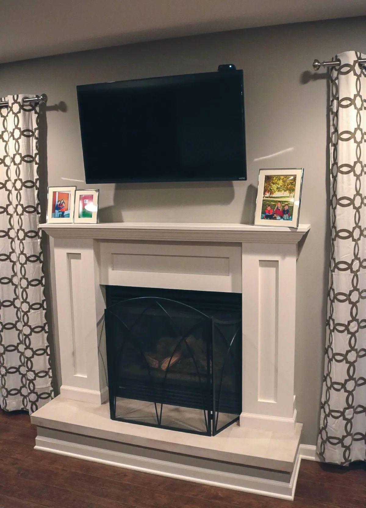 A cozy living room with a prominent white fireplace under a mounted flat-screen tv, flanked by patterned curtains, and decorated with framed photos on the mantel.