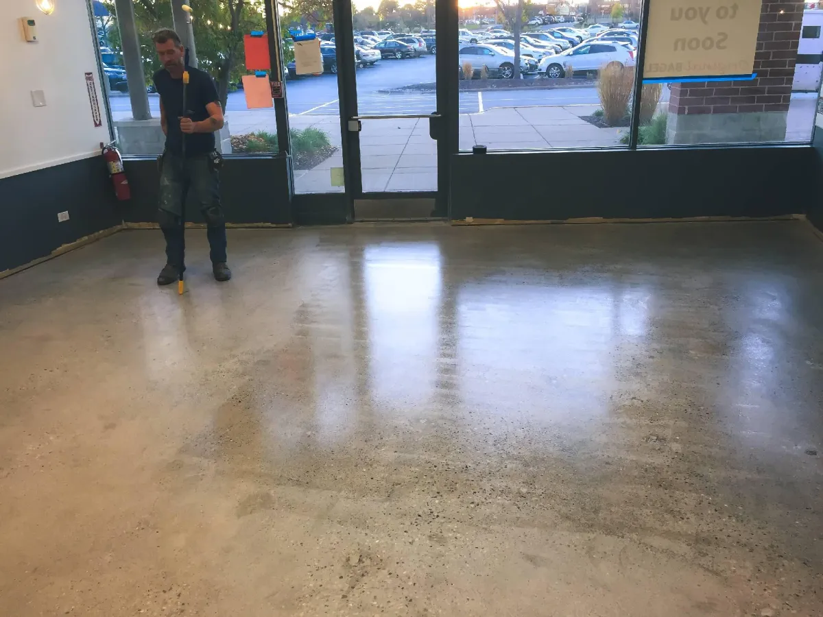 A man stands on a freshly cleaned concrete floor inside a bright room, near large windows showing a parking lot and trees outside, with sunlight casting long shadows indoors.