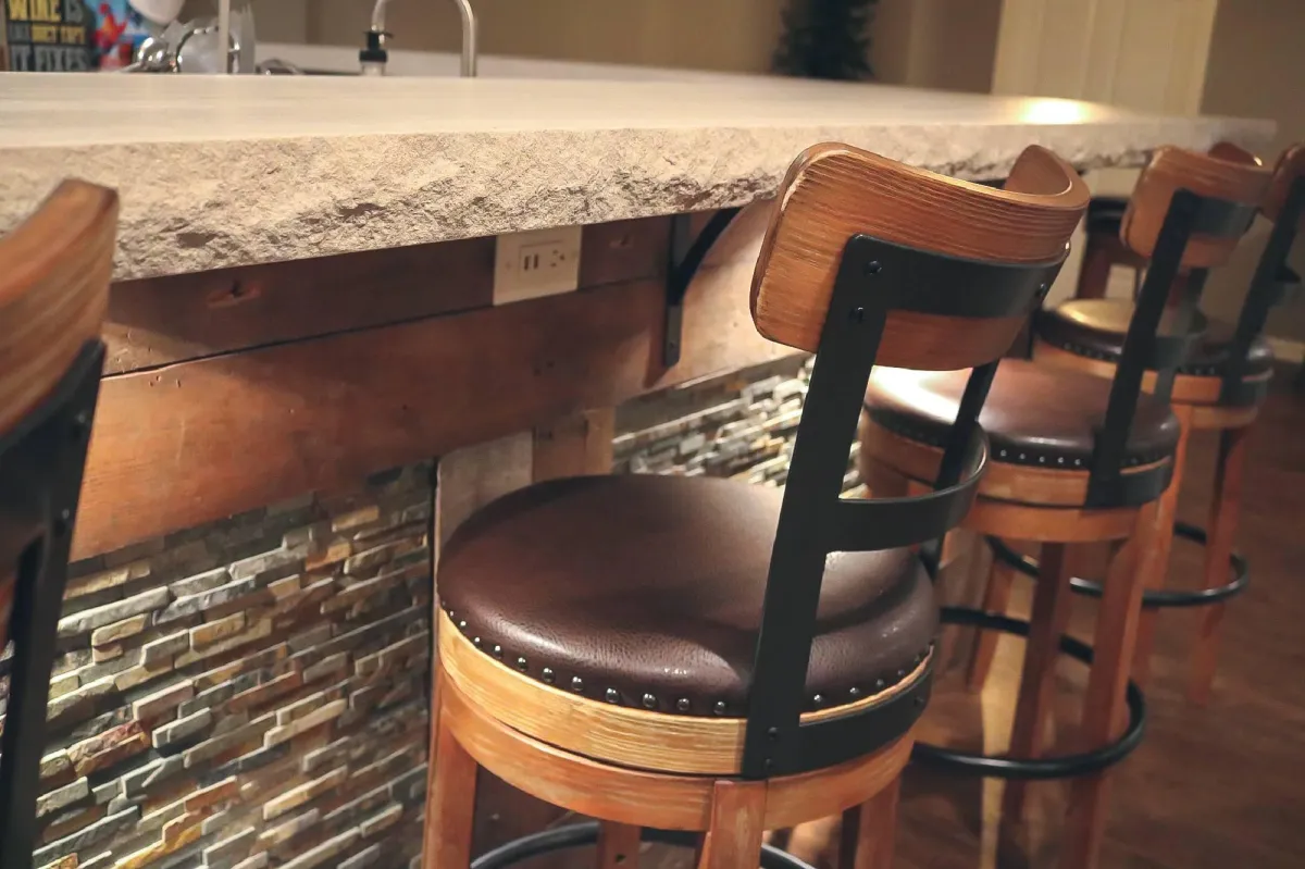 A cozy home bar setup with three high stools with black frames and brown leather cushioning in front of a stone-faced bar counter with a granite top.