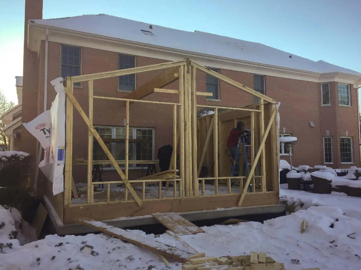 Wooden frame of a small construction project in front of a brick building, with workers on site. snow blankets the ground and rooftops.