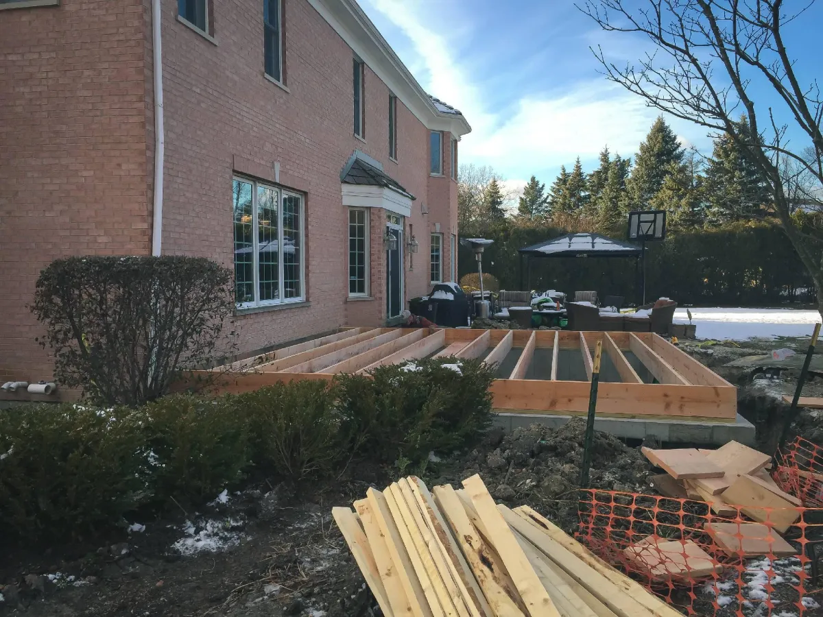 Construction materials and wooden framing for a deck at the side of a brick house, with snowy ground and blue sky in the background.