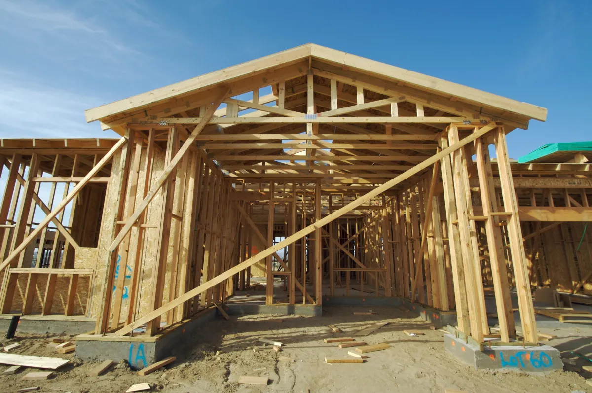 A wooden frame of a house under construction, showing exposed beams and foundations on a clear day.