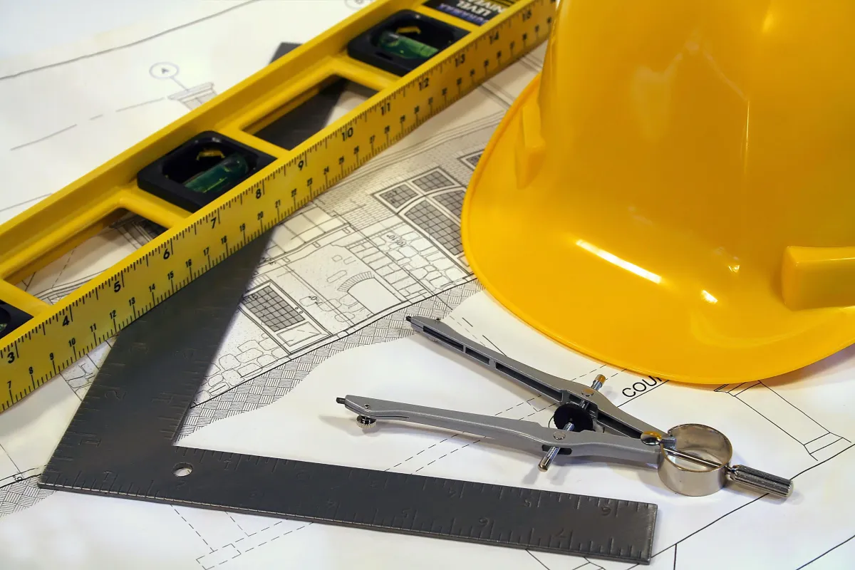 Close-up of architectural blueprints with a yellow hard hat, a folding ruler, and drawing tools including a metal compass and a square on the table.