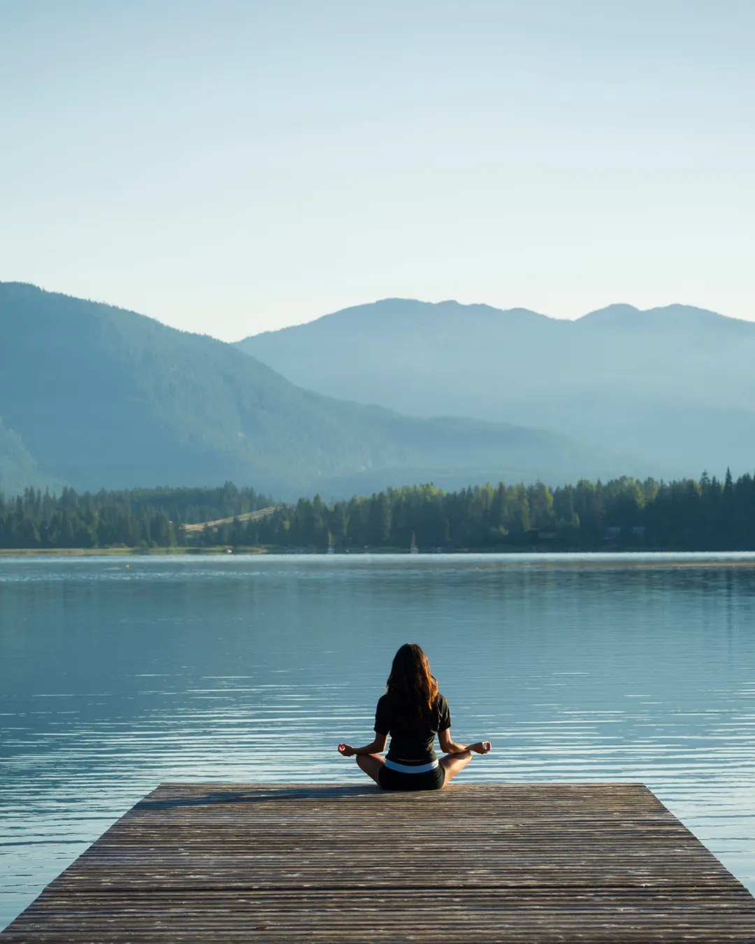 Woman meditating in lotus pose on a wooden dock overlooking a calm mountain lake representing peace and wellness transformation at Solutions Functional Medicine Centre