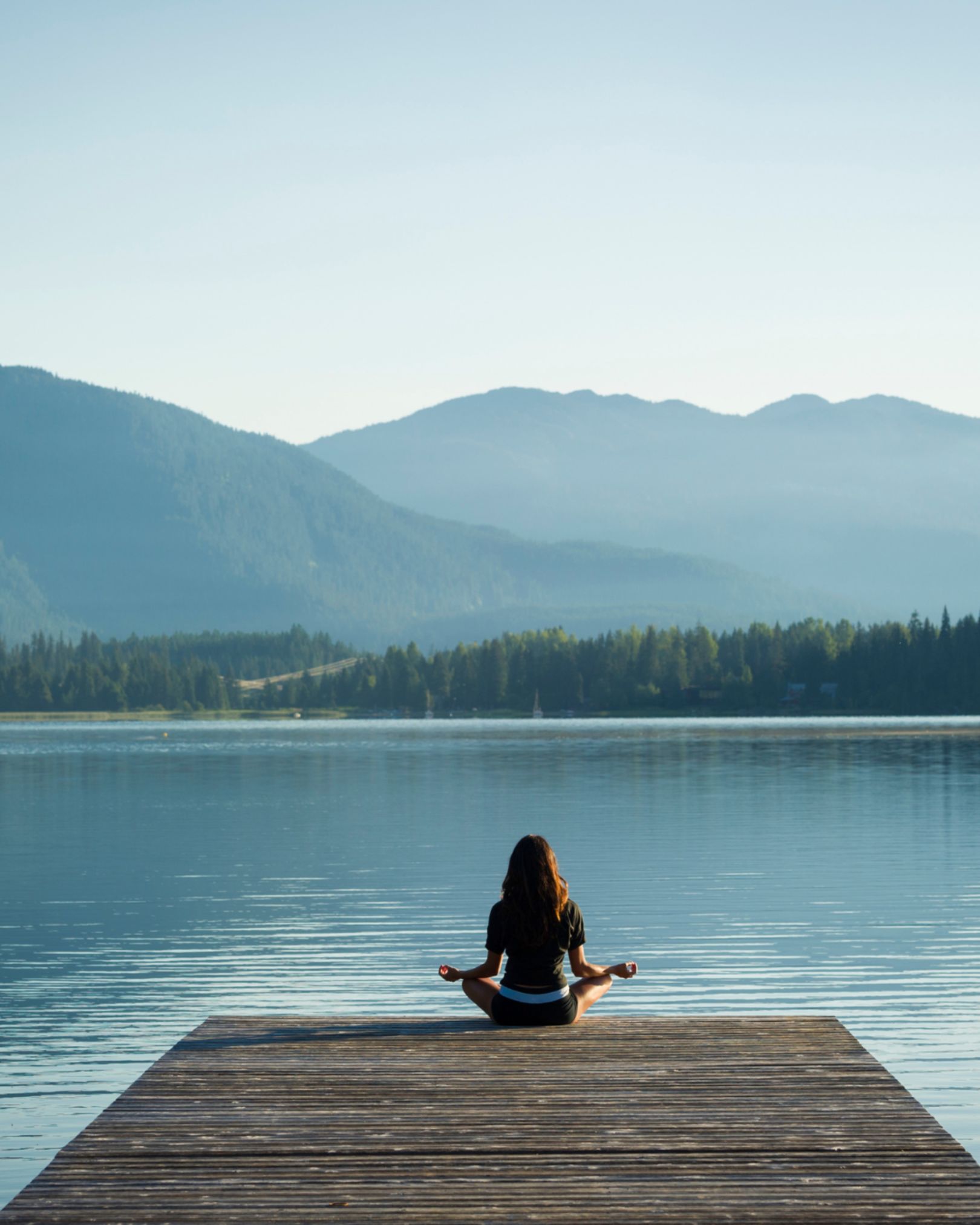 Woman meditating in lotus pose on a wooden dock overlooking a calm mountain lake representing peace and wellness transformation at Solutions Functional Medicine Centre