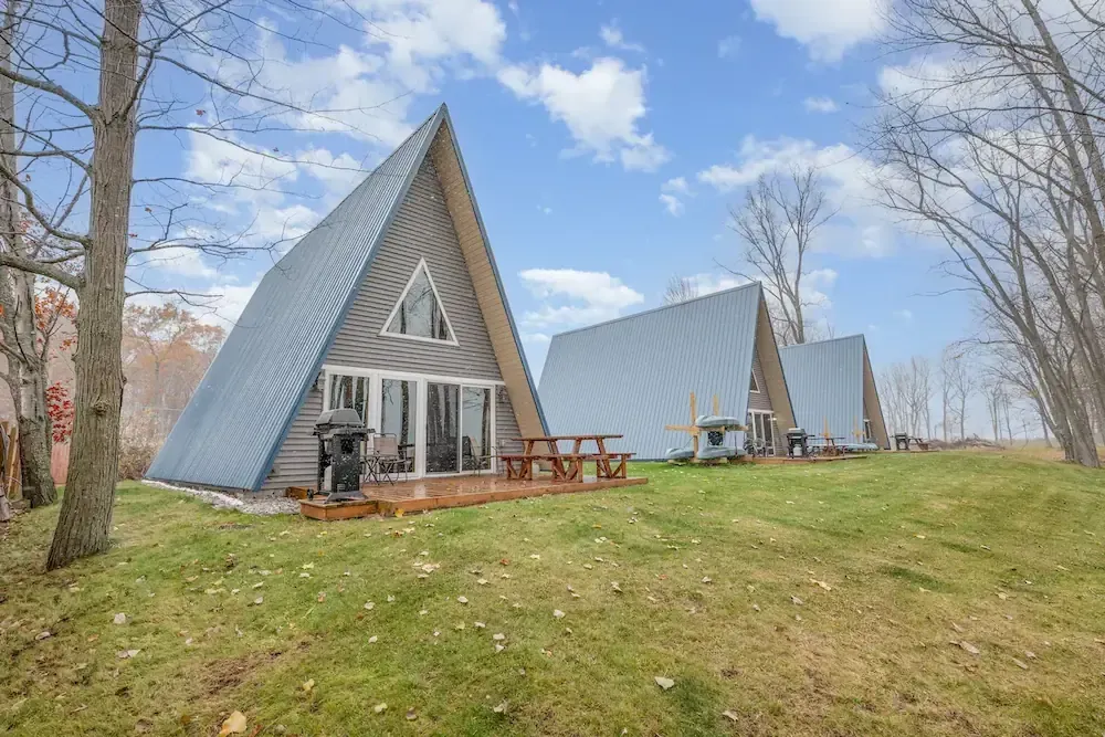 Three modern A-frame cabins sitting side-by-side on a grassy field near Lake Huron, ideal for large group lodging and family reunions.