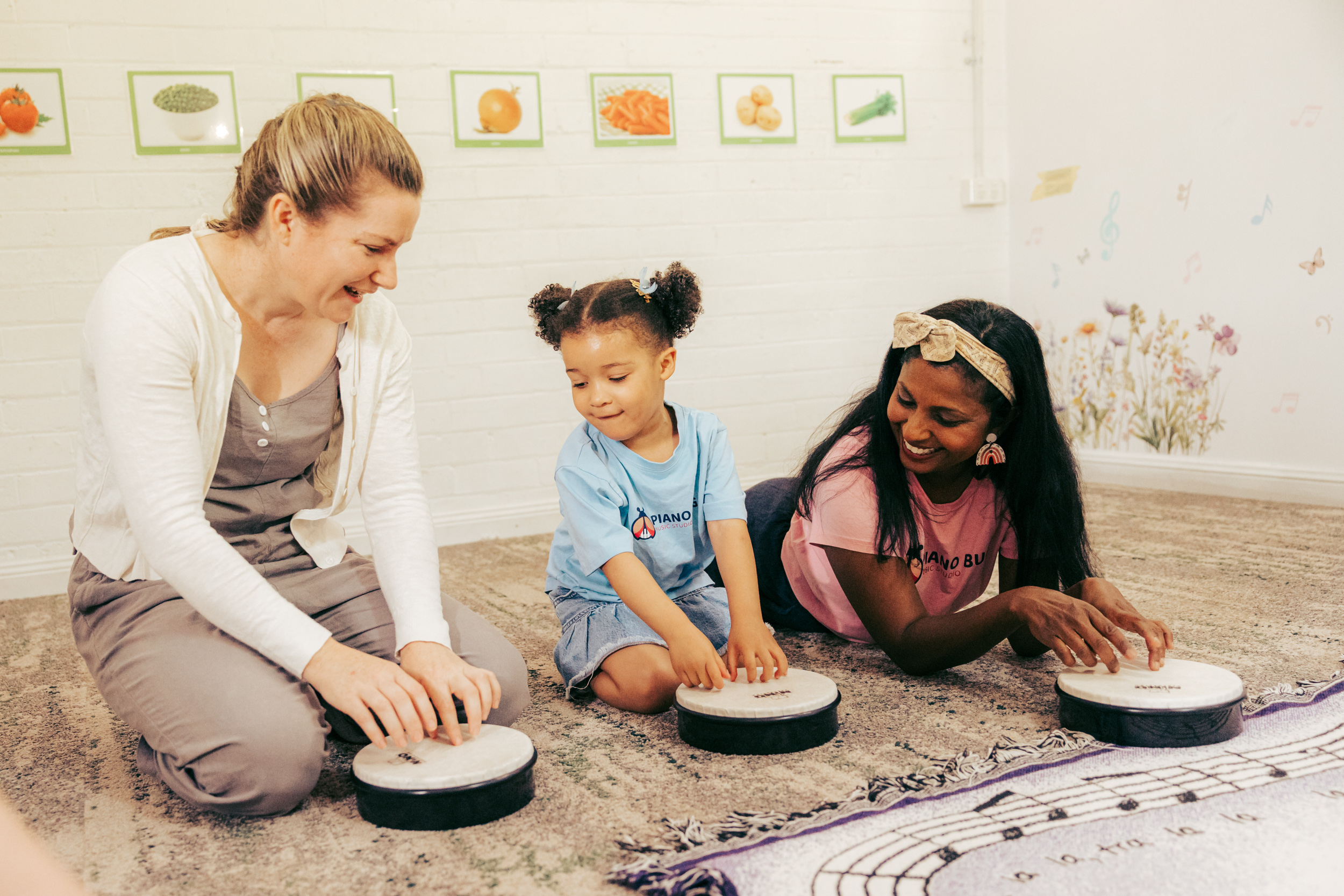 A group of toddlers dance in a circle, holding scarves, while a teacher plays guitar in the background, in a bright, playful classroom with musical decorations.