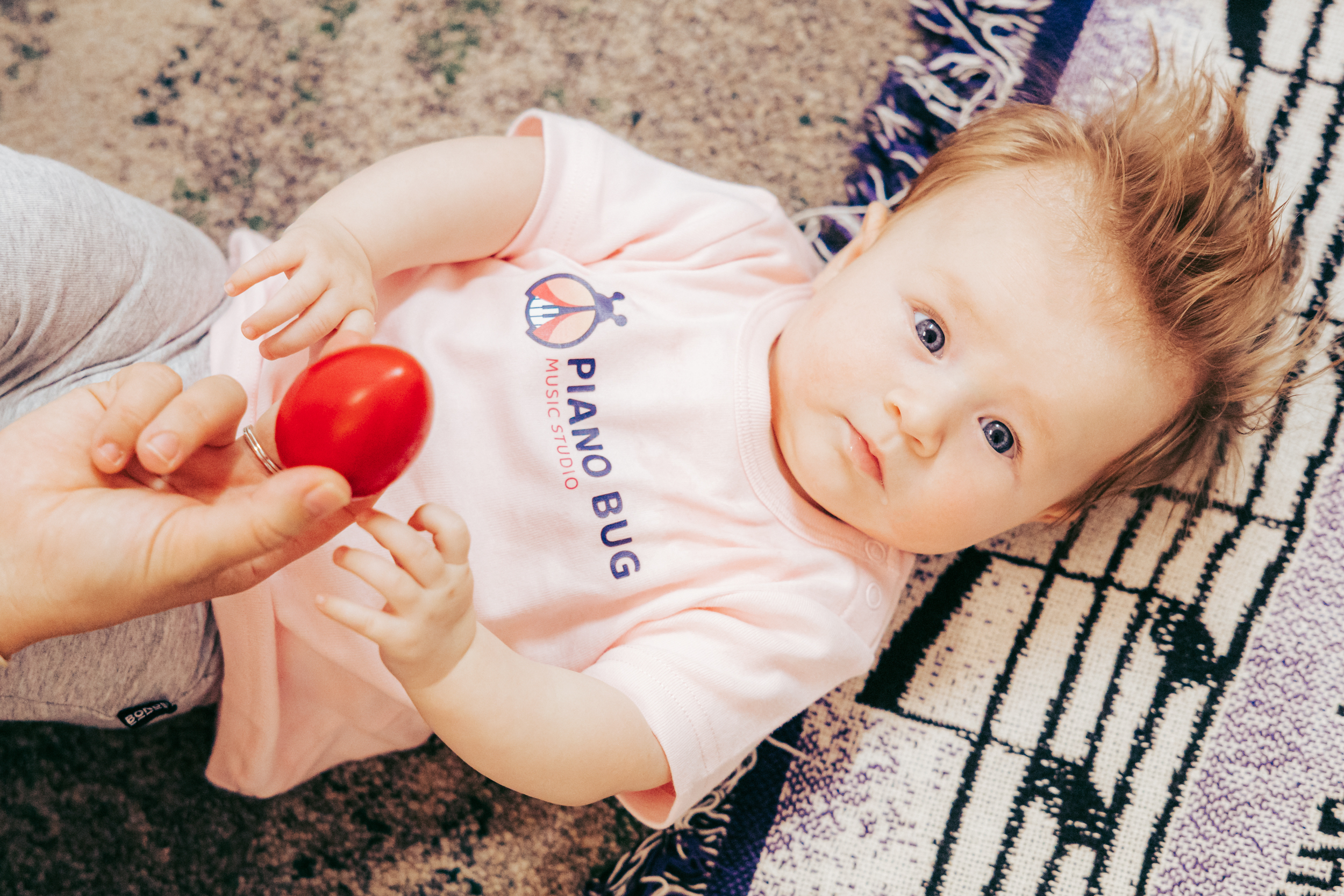 A preschool girl with curly hair laughs as she taps a drum, surrounded by other children and a variety of musical instruments, in a sunlit studio.