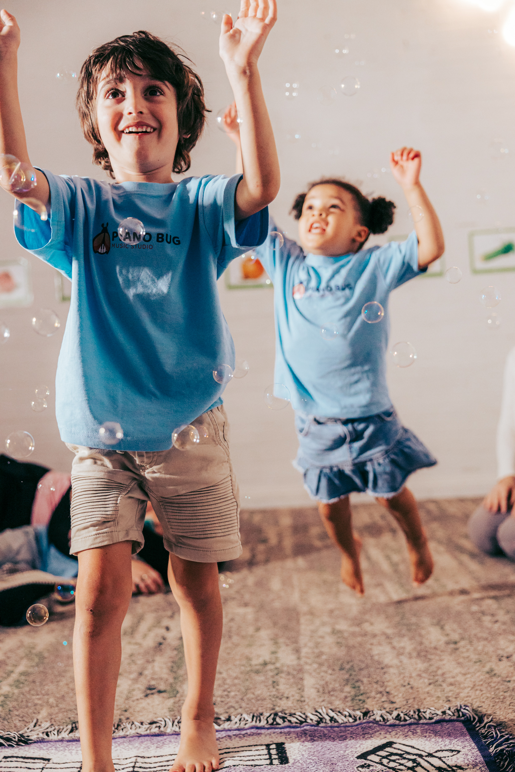 A diverse group of young children and a smiling teacher sit in a circle on a colorful rug, each holding a small instrument, in a bright, welcoming studio with large windows.