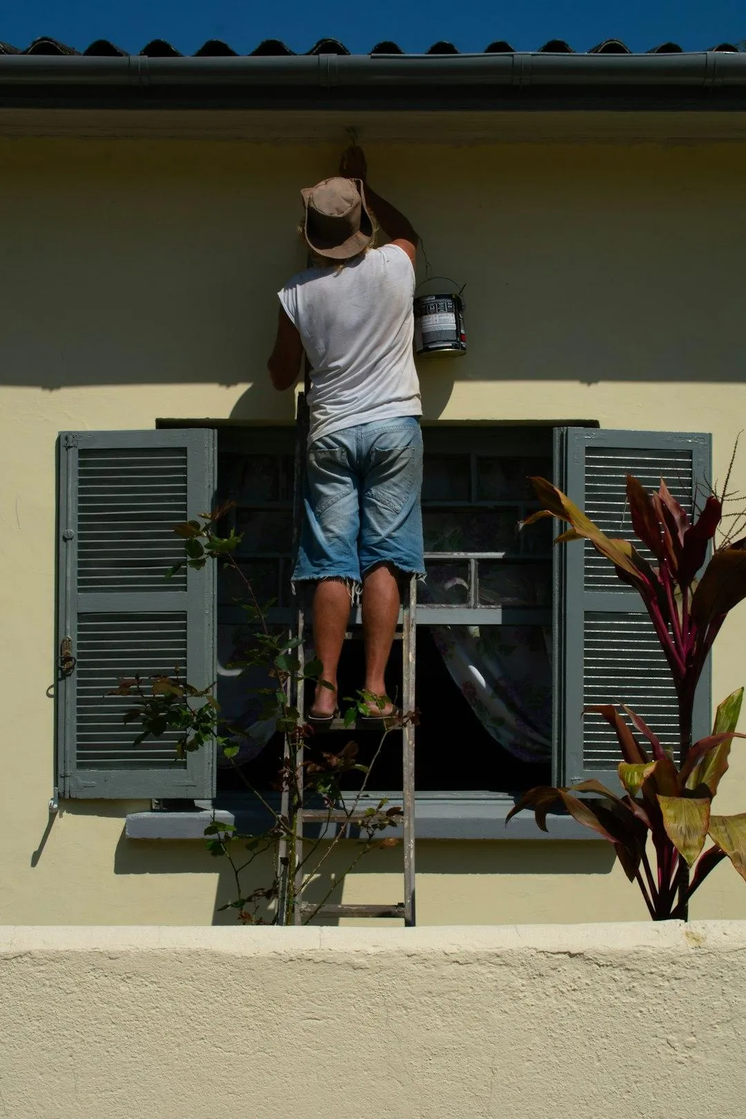 man in singlet and shorts painting house yellow outside window