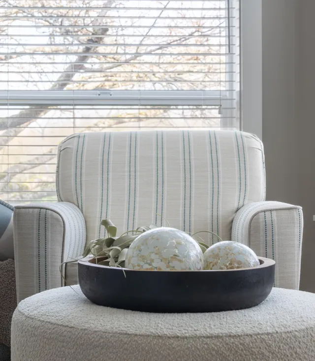  Close-up of a bouclé side table with a dark bowl holding mosaic spheres in front of a striped armchair