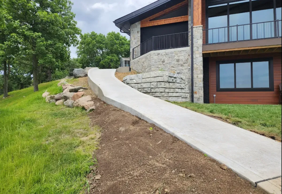 Curved concrete walkway on hillside property next to stone building by Quick Concrete Solutions Iowa
