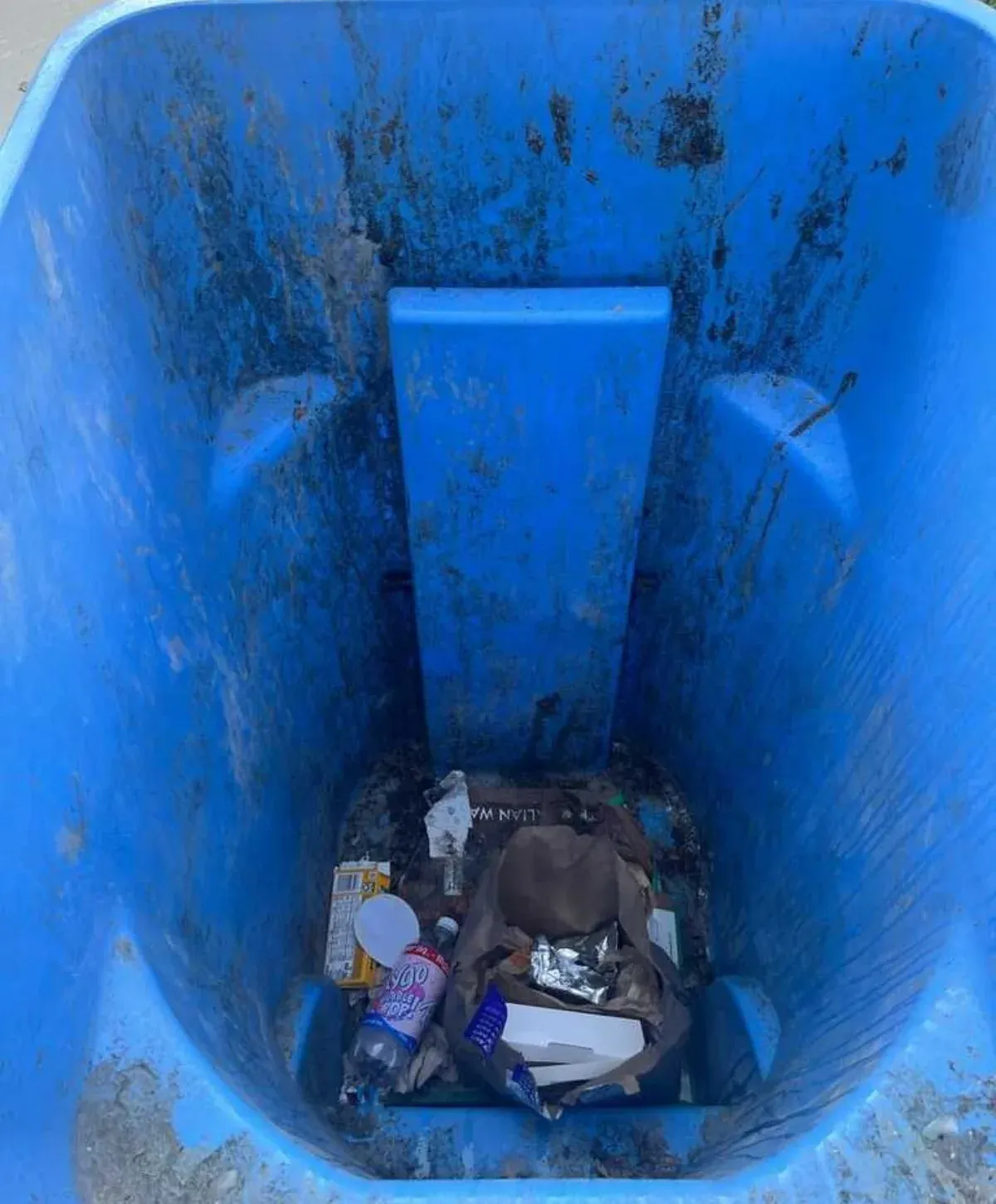An overhead view looking into a dirty blue bin filled with various debris, including a partially visible CrispyRice cereal box.