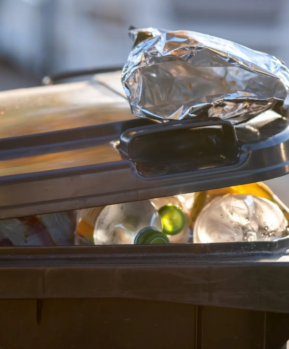 A dark trash bin with its lid slightly open, revealing plastic bottles inside and a crumpled foil bag on top.
