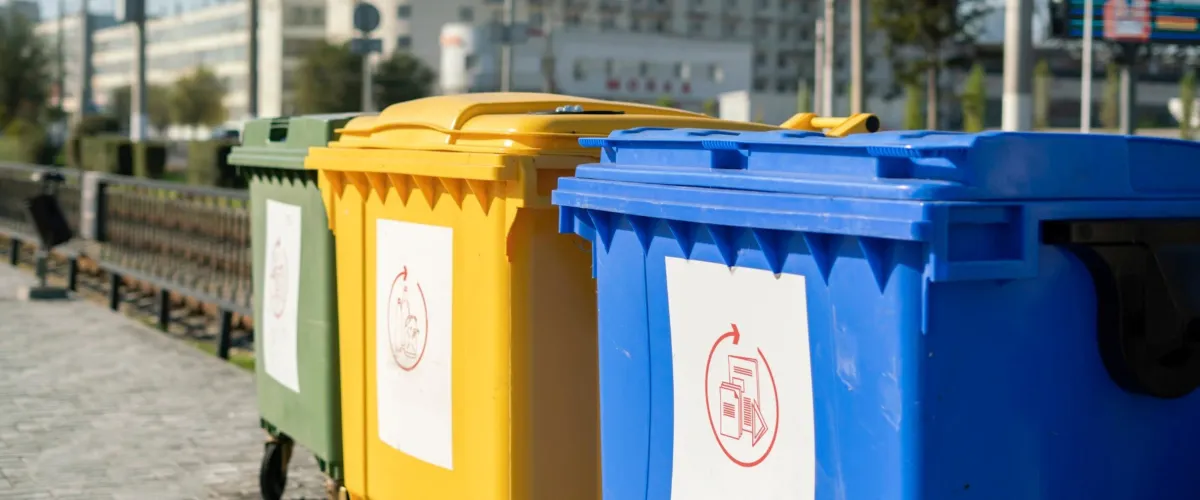 Three brightly colored recycling bins in green, yellow, and blue lined up on a city sidewalk next to a fence.